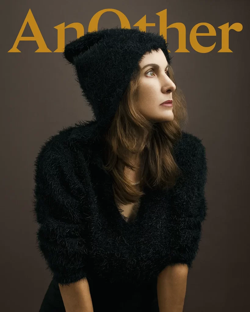 AnOther magazine cover. Woman wearing a fuzzy hooded jacket on a dark background.