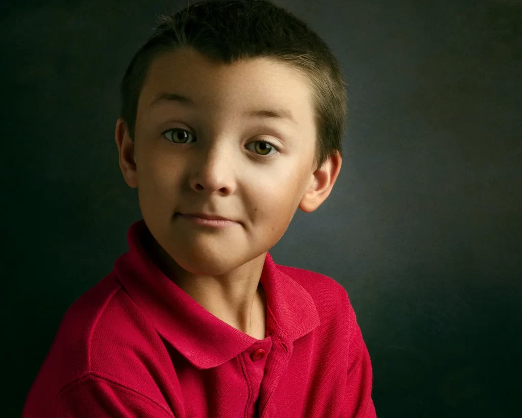 Young boy with a silly expression wearing a red shirt on a dark background.