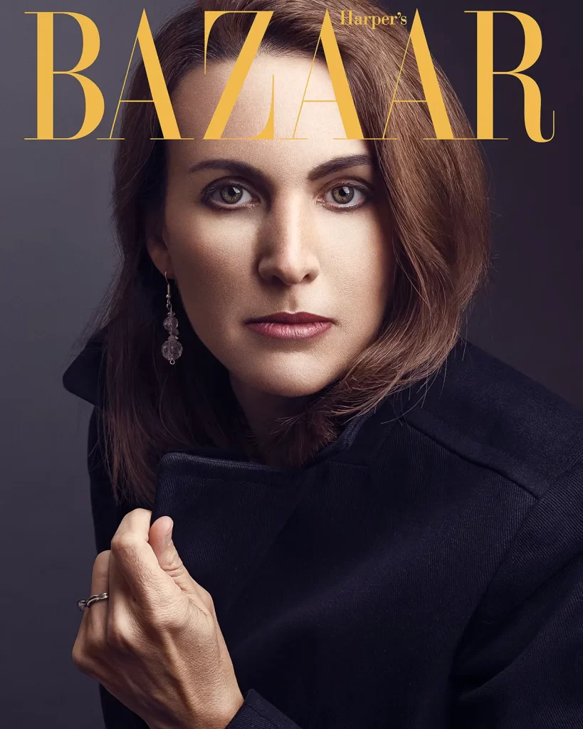 Harper's Bazaar magazine cover. Woman with brunette hair wearing a black coat on a dark background.