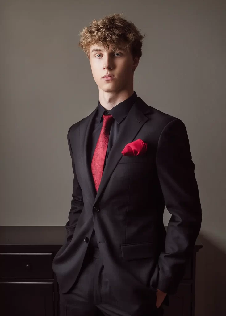 Teenage boy with curly hair wearing a black suit with a red tie.