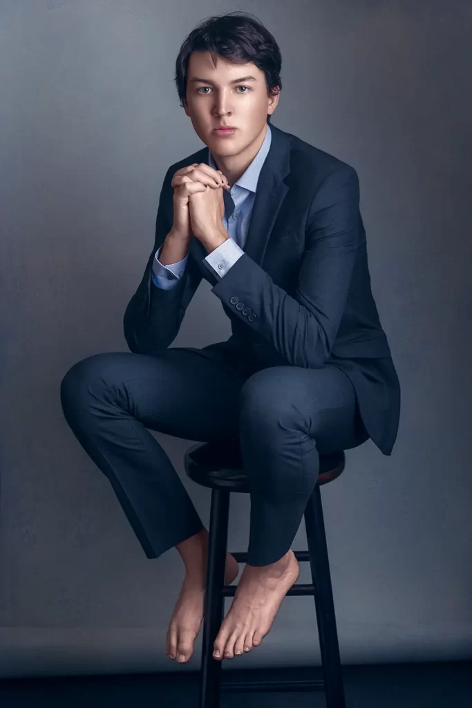 Barefoot young man in a blue suit sitting on a stool.