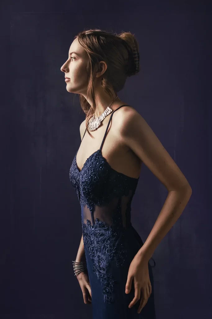 Teenage girl in a blue prom dress with a silver necklace on a dark background.