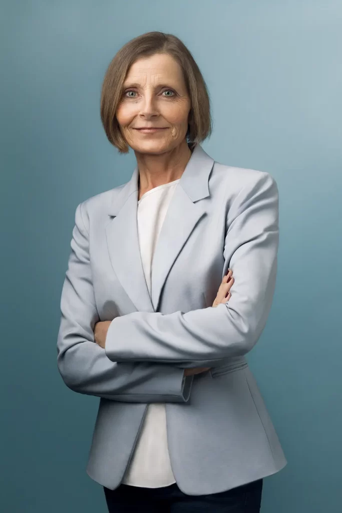 Older business woman with short hair wearing a light blue jacket standing in front of a blue background.