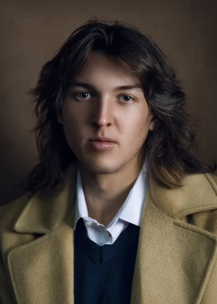 Young man with long dark hair wearing a gold overcoat and white button-down shirt on a brown background.
