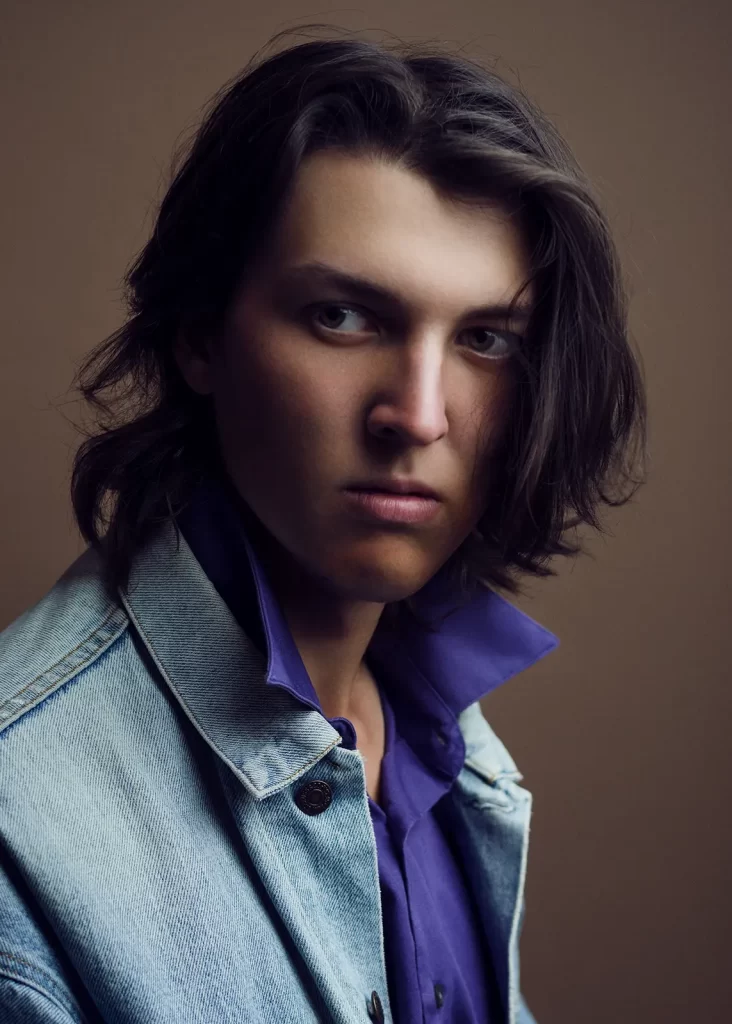 Male college student with long hair in a blue jean jacket and purple button-down on a brown background.