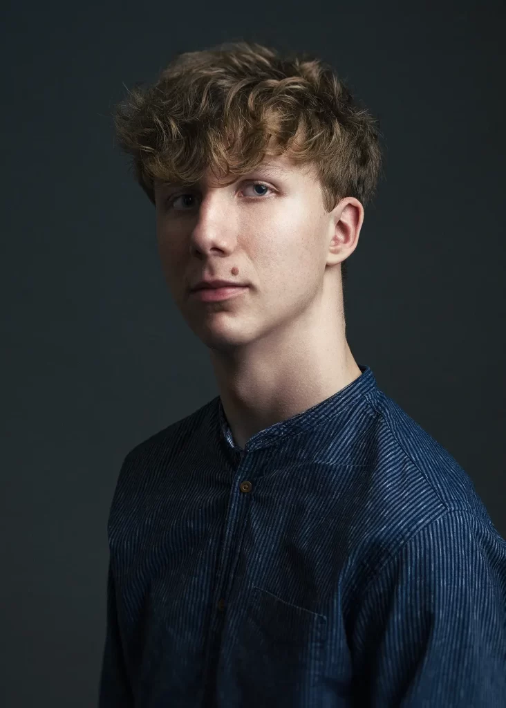 Teenage boy with curly hair in a blue striped shirt on a dark background.