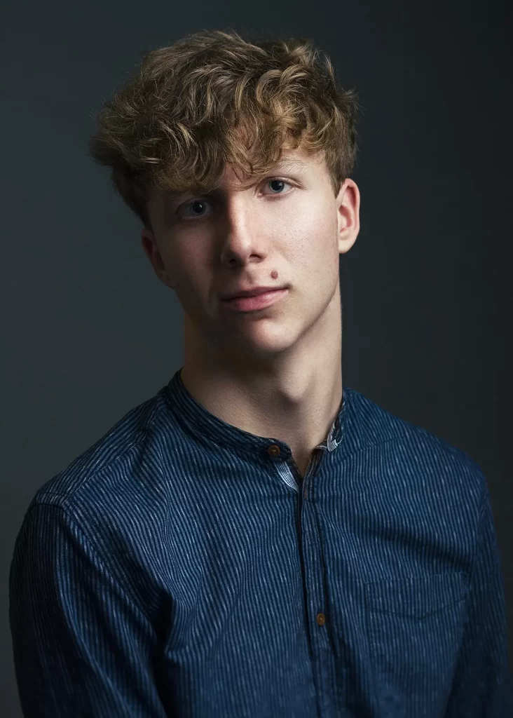 Teenage boy with curly blond hair in a blue striped shirt on a dark background.