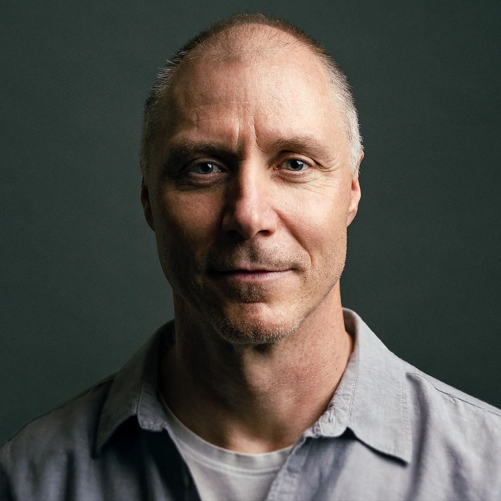 Middle-aged man with short hair and face stubble wearing a gray shirt on a dark background.