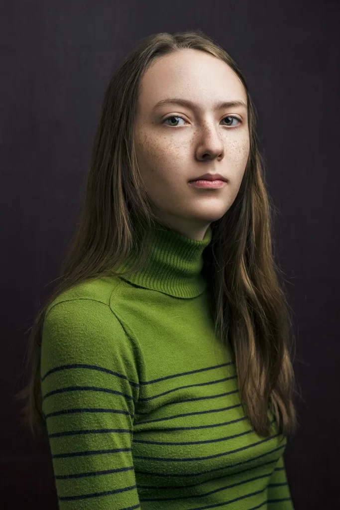 Teenage girl with long straight hair wearing a green striped sweater on dark background.