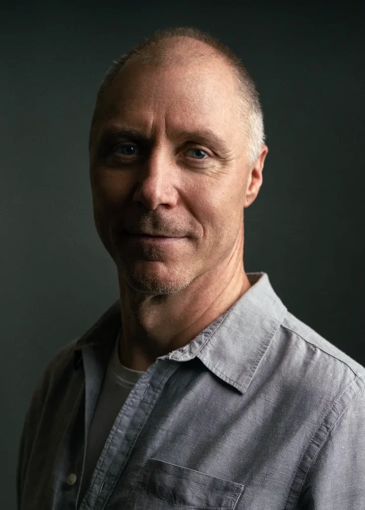 Balding middle-aged man with short hair and face stubble wearing gray shirt.