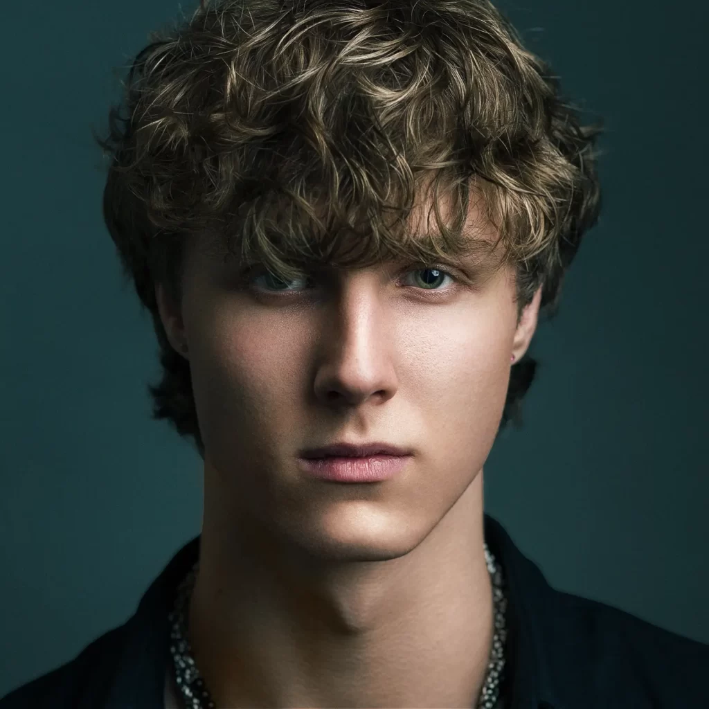 Close-up portrait of a teenage boy with curly hair in a black shirt on a dark green background.