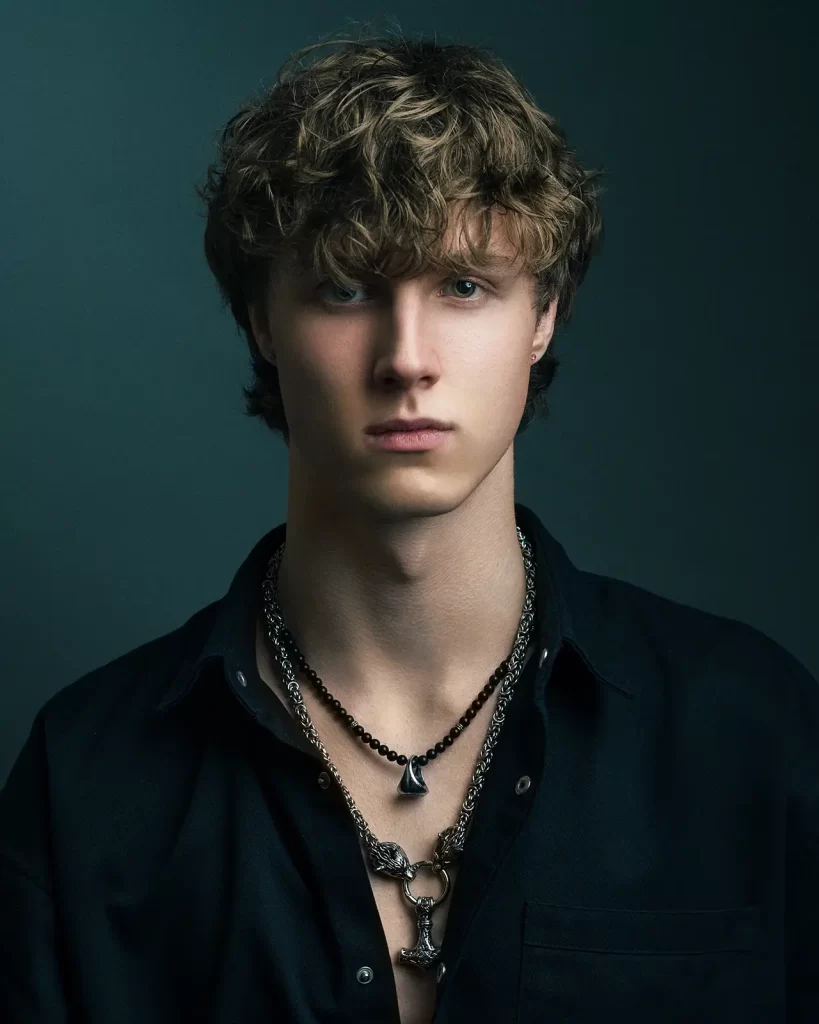 Teenage boy with curly hair wearing a black shirt and two necklaces on a dark green background.