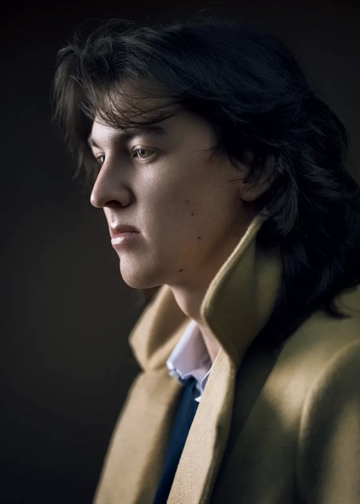 Young man with long hair wearing a gold overcoat with a popped collar on a dark brown background.