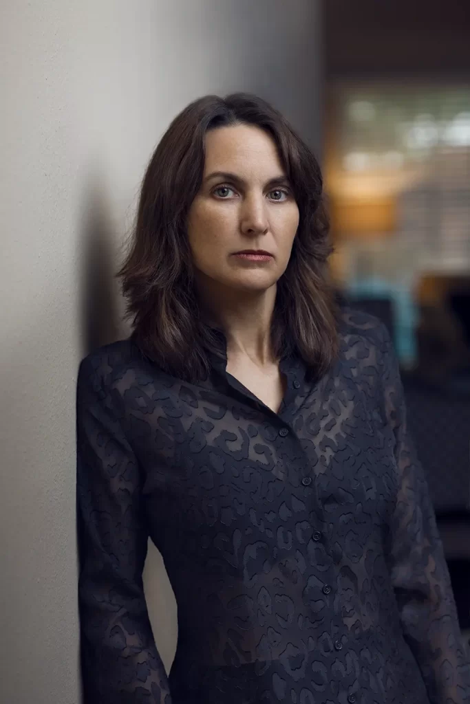 Attractive brunette woman with long hair leaning against a wall wearing a black textured blouse.