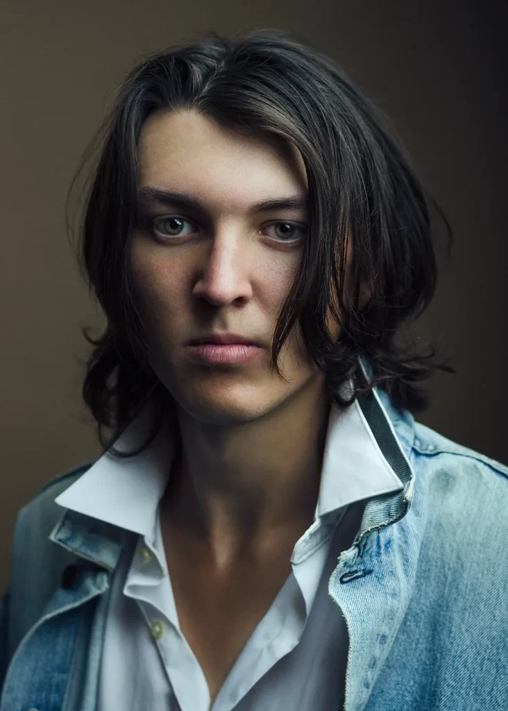 Male college student with long hair in a blue jean jacket on a brown background.