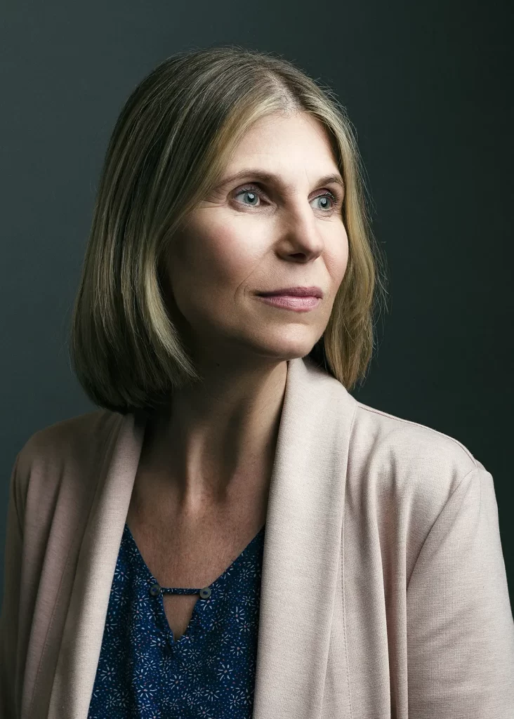Middle-aged woman with blonde hair wearing a tan sweater on a dark background.
