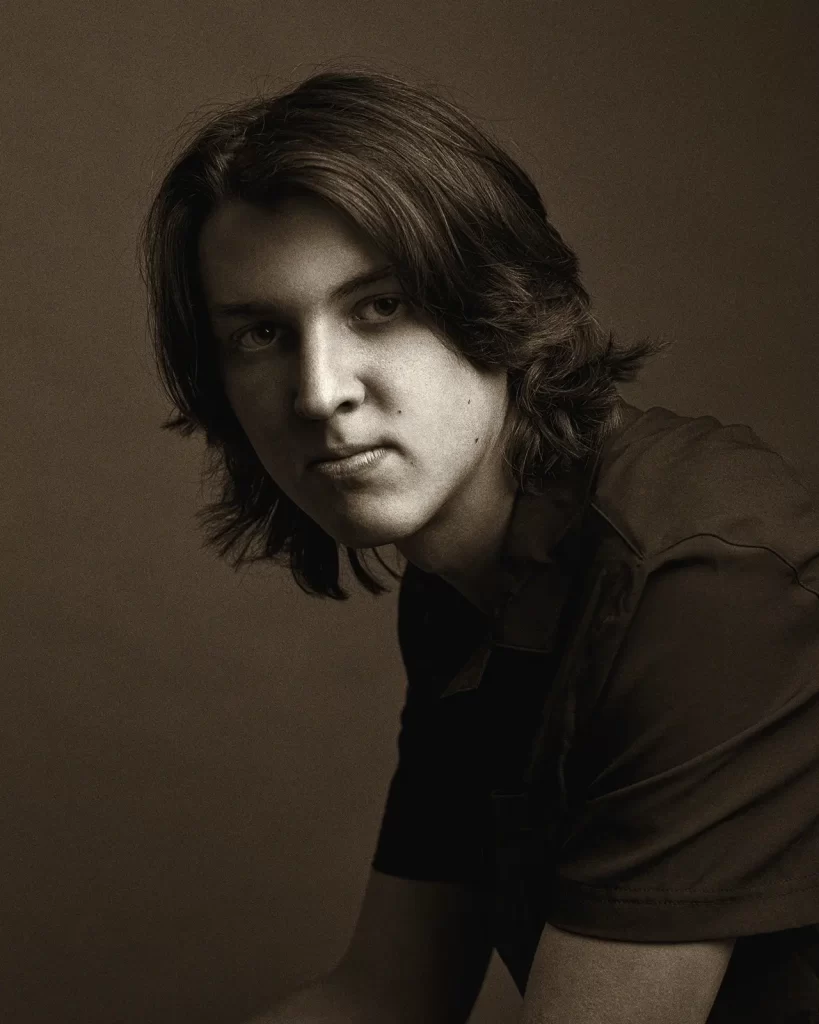 Warm black and white portrait of a teenage boy with long dark hair on a dark background.