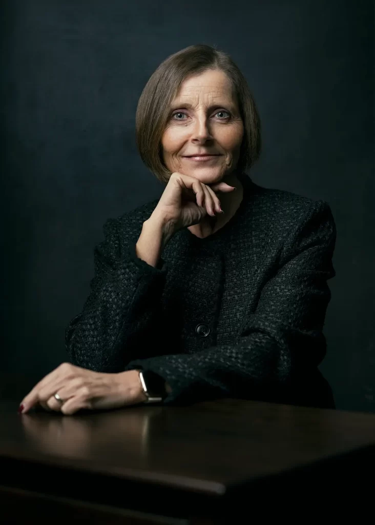Older business woman with short hair wearing a black jacket sitting in front of a dark textured background.