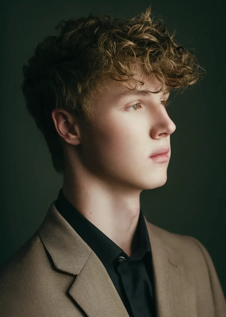 Teenage boy with curly hair wearing a tan suit jacket and a black shirt.