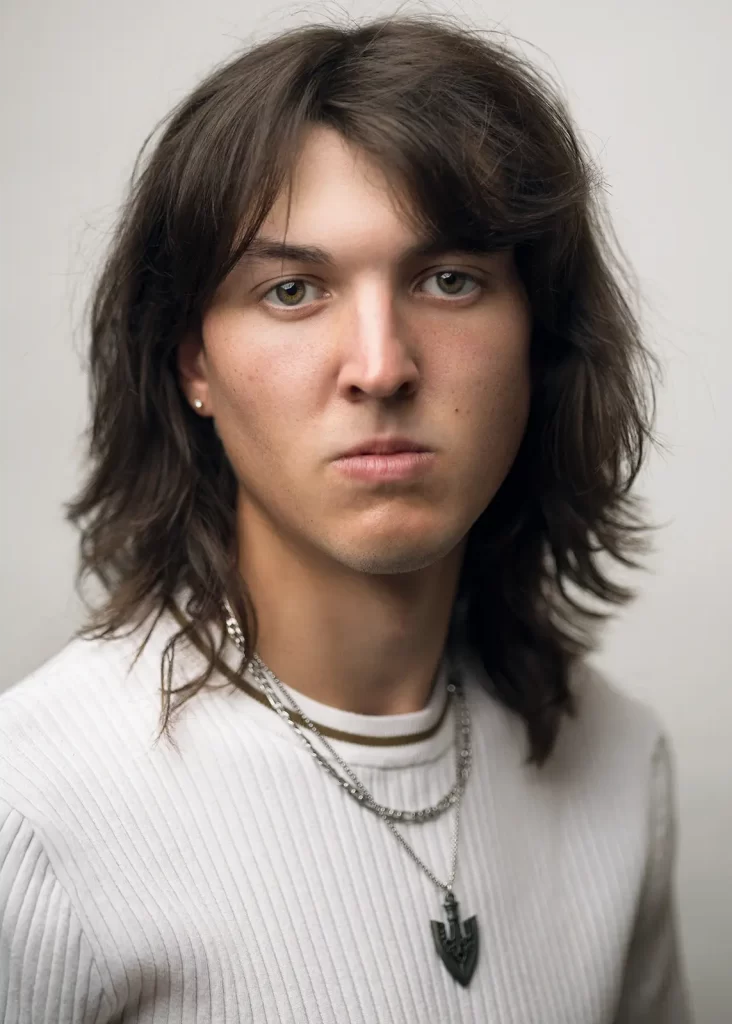 Young man with long hair and an earring wearing a white ribbed shirt in front of a white background.