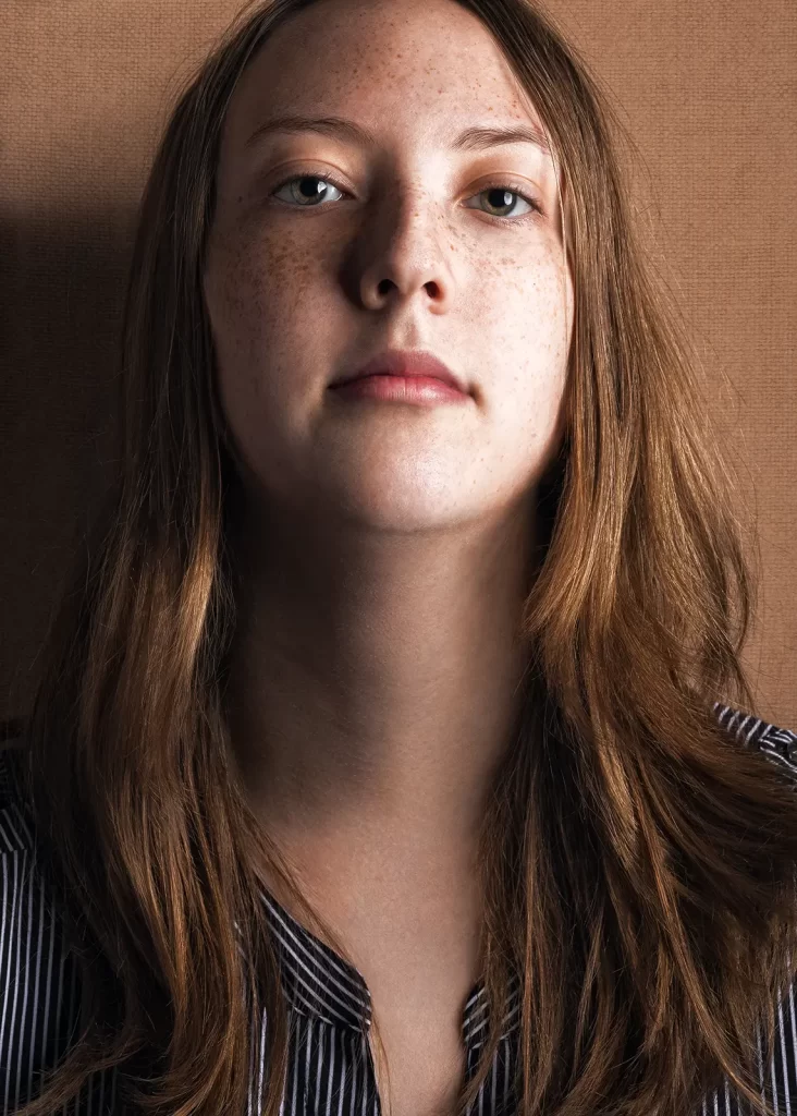 Young woman with long sandy blonde hair and an attitude leaning against textured brown background.