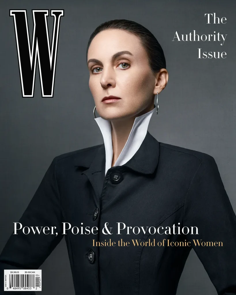 W magazine cover. Woman with slicked back hair wearing a black jacket and white blouse with an extended collar on a dark gray background.